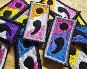 May include: A close-up of a pile of embroidered patches featuring the semicolon symbol in various colors, including yellow, purple, blue, pink, and white. The patches are made of fabric and have a black embroidered semicolon on each one.