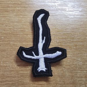 Puede incluir: Parche bordado en blanco y negro con un diseño de cruz invertida estilizada. La cruz tiene forma de árbol. El parche está diseñado para coserse en ropa o accesorios.