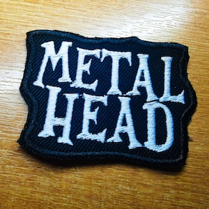 May include: A black fabric patch with white embroidered text that reads "METAL HEAD".