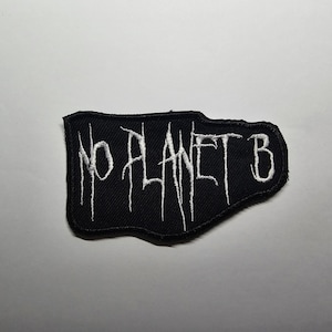 May include: Black embroidered patch with the words "NO PLANET B" in white, stylized lettering. The patch has a jagged, irregular shape and is likely designed to be sewn onto clothing or accessories.