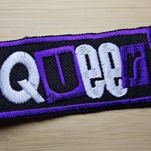 May include: Embroidered patch with the word "QUEER" in white and purple thread. The patch has a black background with a purple border. The patch is rectangular and appears to be made of fabric.