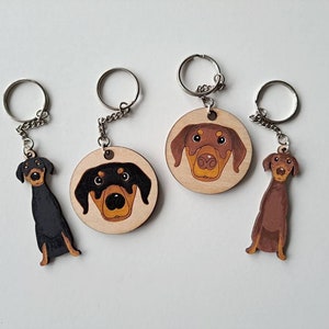 May include: Three wooden keyrings featuring different dog breeds. Each keyring has a round wooden charm with a painted dog face and a silver metal key ring. The dog breeds are a Doberman Pinscher, a Dachshund, and a Labrador Retriever.