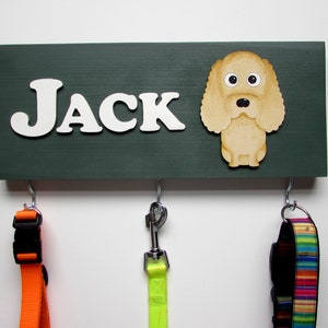 Könnte beinhalten: Ein dunkelgrüner, hölzerner Hunde-Leinenhalter mit dem Namen "JACK" in weißen Buchstaben. Ein Cartoon-Hund befindet sich auf der rechten Seite des Halters. Es gibt drei Haken zum Aufhängen von Leinen. Zwei Leinen hängen an den Haken, eine orange und eine gelbe.