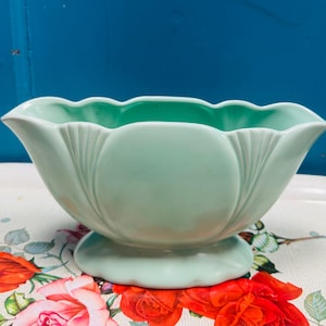 May include: A pale green ceramic bowl with a scalloped edge and a pedestal base. The bowl has a flower-like form with vertical ridges. This vintage bowl is a decorative item, likely for display.