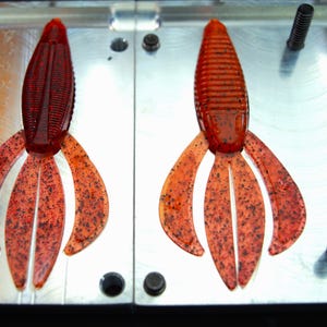 May include: Two red and orange soft plastic fishing lures with a speckled finish. The lures are in a mold with the text "LURECRAFT 5XL-U-178" visible.