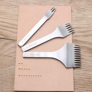 May include: Three silver leather stitching tools with different sized prongs. The tools are laid out on a light brown leather surface. The tools are labelled with the measurement 3.85mm.
