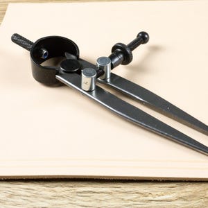 Puede incluir: Un compás negro y plateado, utilizado para medir y dibujar círculos, descansa sobre una superficie de cuero de color beige claro. El compás tiene dos patas puntiagudas, un mecanismo de ajuste central y un mango negro. La herramienta está diseñada para la precisión.