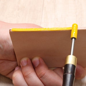May include: A person is using a tool to apply a yellow adhesive to the edge of a piece of leather.