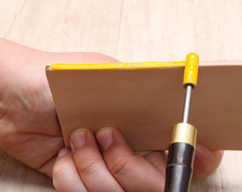 May include: A person is using a tool to apply a yellow adhesive to the edge of a piece of leather.