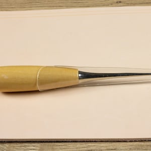 Puede incluir: Un punzón de costura de cuero de madera y metal con una cubierta protectora de plástico transparente. El punzón se utiliza para perforar agujeros en el cuero para coser.