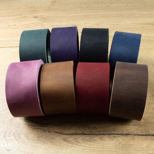 May include: A collection of eight leather cuffs in a range of colours: green, purple, black, blue, pink, brown, red, and dark brown. The cuffs are displayed on a wooden surface.