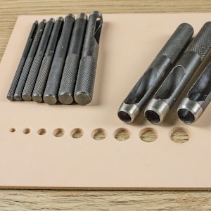 Puede incluir: Una colección de herramientas de marroquinería, que incluye punzones y un juego de punzones de varios tamaños, dispuestos sobre una superficie de cuero de color beige claro. Las herramientas son de metal oscuro con mangos texturizados.