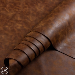 May include: A roll of medium brown leather featuring a mottled, variegated surface texture with darker and lighter brown tones throughout. The material is flexible and shows a smooth, matte finish with a visible grain pattern across the surface.