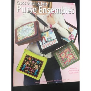 May include: A book cover titled "Cross Stitch & Felt Purse Ensembles" featuring various cross-stitched purse designs. The purses showcase diverse patterns, including floral, mermaid, and shopping-themed motifs. The cover is pink with the text "Cross Stitch & Felt Purse Ensembles".