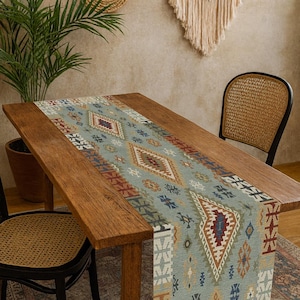 Può includere: Runner da tavolo rettangolare con motivo geometrico nei toni del blu, rosso, marrone e crema. Il runner è posto su un tavolo di legno. La stanza ha uno stile bohémien con una sedia intrecciata e decorazioni murali.