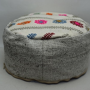 May include: A round, white and grey ottoman with a colourful embroidered top. The ottoman has a woven texture and is perfect for adding a touch of bohemian style to any room.