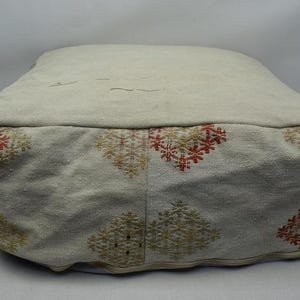 May include: A square, beige pouf with embroidered floral patterns in red, yellow, and green. The pouf is made of a textured fabric and has a soft, cushioned appearance, suitable for seating or as a decorative accent.