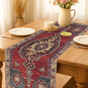 May include: A patterned table runner in a traditional rug style. The runner has a deep red central field with beige, grey, and blue patterns. It is placed on a wooden table, with a vase of flowers and tableware.