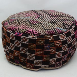 May include: A round, black and white patterned ottoman with pink and blue accents. The ottoman is made of woven fabric and has a slightly rounded top.