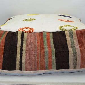 May include: A square, floor cushion with a white top and a striped base. The top features colorful geometric shapes. The base has vertical stripes in shades of brown, orange, and green. The cushion is approximately 24 inches wide and 12 inches tall.