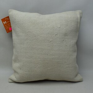 Può includere: Un cuscino quadrato tessuto bianco sporco. Il cuscino ha una superficie strutturata e un'etichetta attaccata al lato. Il cuscino è di colore neutro e completerebbe vari stili di arredamento.