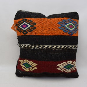 May include: A black, orange, red, and white patterned pillow with geometric designs. The pillow is made of woven fabric and has a fringed edge.