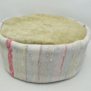 May include: Circular ottoman with a beige top and a woven base in various colours. The base has vertical stripes of pink, blue, and yellow. This decorative piece is suitable for living rooms or bedrooms.