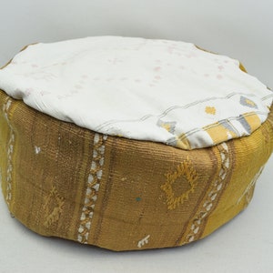 May include: A round, brown and white patterned pouf with a white fabric top. The pouf is made of woven fabric and has a traditional design.