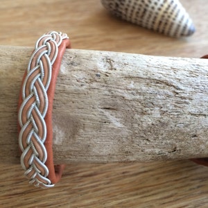 Könnte beinhalten: Ein geflochtenes Lederarmband mit einem silbernen Draht, der in das Design eingewebt ist. Das Armband ist braun und hat einen natürlichen, rustikalen Look.