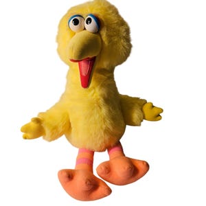 May include: A plush yellow bird character with large white eyes, a yellow beak, and a red tongue. The bird has orange feet and legs with pink and orange stripes. The arms are outstretched, and the bird appears to be smiling.