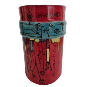 May include: A handmade ceramic tumbler with a vibrant red base, adorned with teal and yellow accents. The tumbler features black line art and a teal band around the middle. The design includes abstract shapes and patterns.