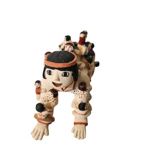 May include: A handcrafted ceramic figurine depicting a person carrying several smaller figures. The main figure has black hair, an orange headband, and a smiling face. The smaller figures are wearing hats and various colors. The figurine is off-white with painted details.