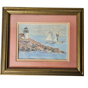 May include: Framed artwork featuring a coastal scene. The pastel drawing depicts a white lighthouse and a small boat sailing on blue water. The artwork is set against a pink mat and framed in a gold-colored frame.