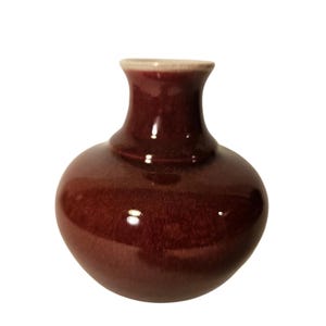 May include: A small, round, maroon-colored ceramic vase with a narrow neck and a slightly flared rim. The vase has a glossy finish and a smooth, rounded body. The rim is a lighter, beige color.