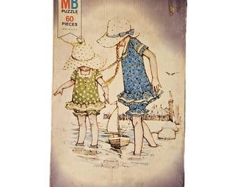 May include: Vintage MB puzzle box featuring an illustration of two children in dresses and sun hats playing near the water. The box reads "MB PUZZLE 60 PIECES MADE IN U.S.A." The artwork is in soft pastel colors.