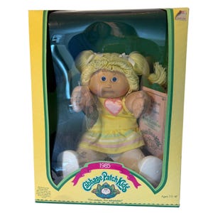 May include: A 1985 Cabbage Patch Kids doll with blonde yarn hair in pigtails, blue eyes, and a yellow dress featuring a pink heart. The doll is packaged in a yellow box with the text Cabbage Patch Kids, 1985, and I'm unique. I'm adoptable.