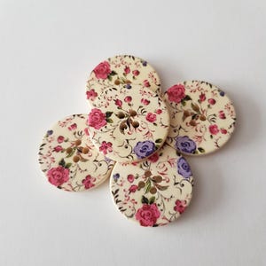 Könnte beinhalten: Fünf runde Holzknöpfe mit floralem Design. Die Knöpfe haben einen cremefarbenen Hintergrund mit rosa und lila Rosen und grünen Blättern. Jeder Knopf hat vier Löcher zum Nähen.