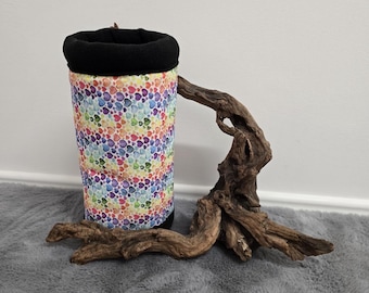 May include: A cylindrical drinks sleeve with a black rim and a colourful heart pattern. It rests on a piece of driftwood. The background is light grey.