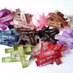Könnte beinhalten: Sortiment an bunten Stoff-Etiketten mit dem aufgedruckten Text "HAND MADE". Die Etiketten sind in verschiedenen Farben, darunter Rot, Blau, Grün, Lila und Braun.