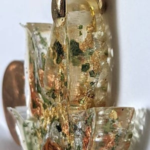 May include: Gold pendant with a clear resin design featuring green, gold, and copper accents. The resin design is reminiscent of a flower or a leaf.
