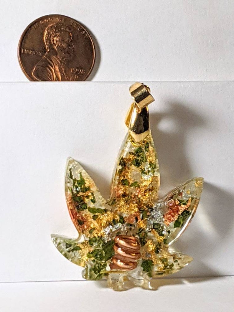 May include: Gold-tone pendant featuring a clear resin leaf with embedded green and gold flakes, copper wire, and a gold-tone bail.