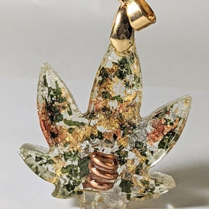 May include: A gold-tone pendant shaped like a marijuana leaf with green, gold, and copper accents. The pendant is made of clear resin with embedded metallic flakes and a copper coil.