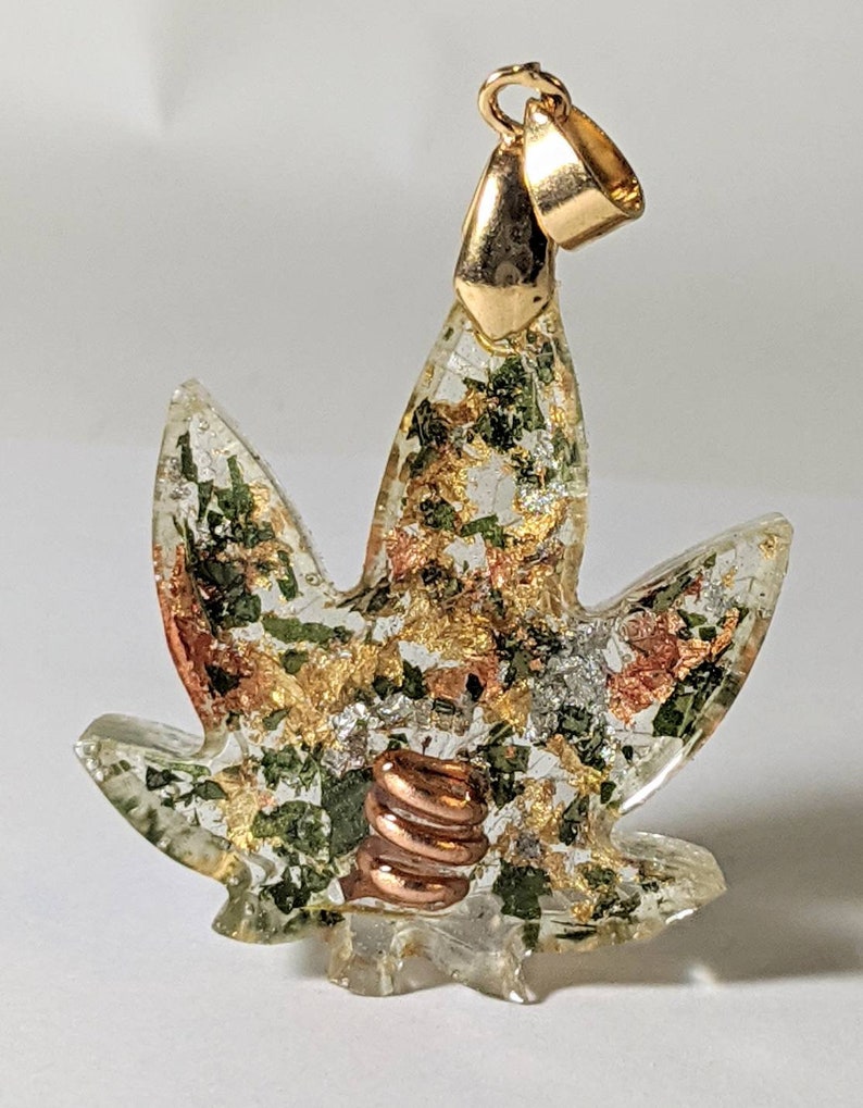 May include: A gold-tone pendant shaped like a marijuana leaf with green, gold, and copper accents. The pendant is made of clear resin with embedded metallic flakes and a copper coil.