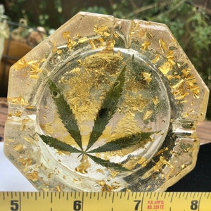May include: Clear resin ashtray with a cannabis leaf and gold flakes embedded inside. The ashtray has an octagonal shape and is approximately 7 inches in diameter. The ashtray is a decorative item.
