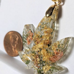 May include: Gold-tone pendant featuring a clear resin leaf with embedded gold and green flakes. The pendant is attached to a gold-tone bail.