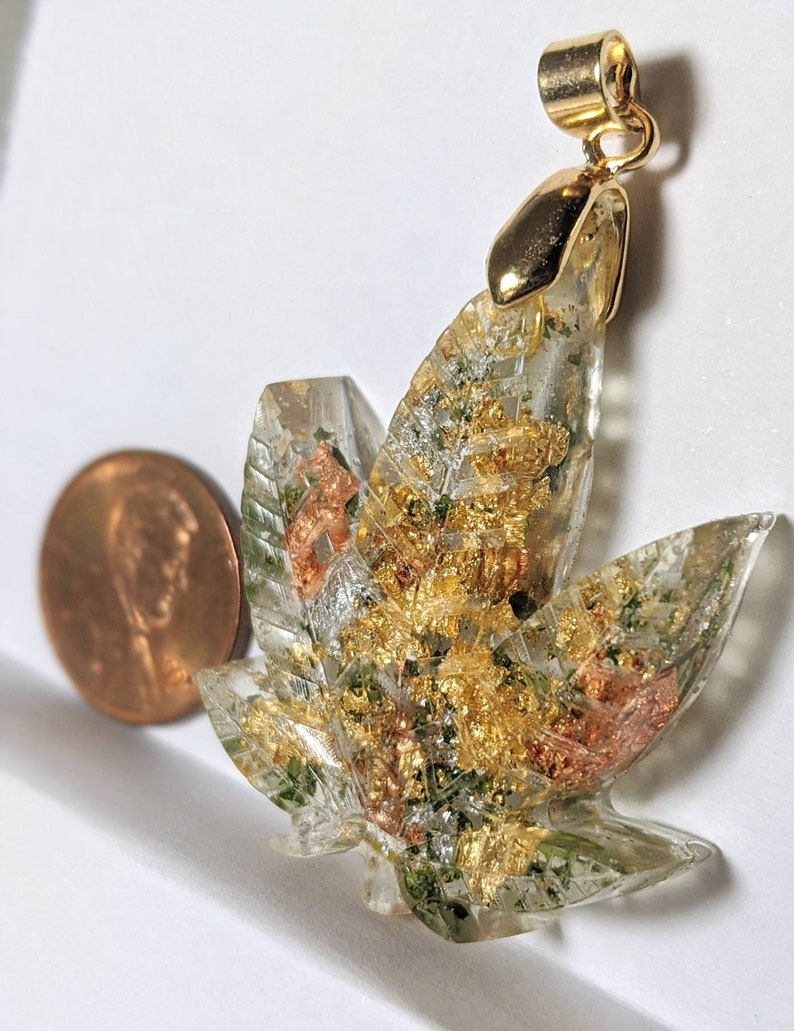 May include: Gold-tone pendant featuring a clear resin leaf with embedded gold and green flakes. The pendant is attached to a gold-tone bail.
