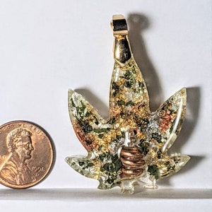 May include: Gold-tone pendant in the shape of a marijuana leaf with green, gold, and copper accents. The pendant is approximately the size of a US penny.