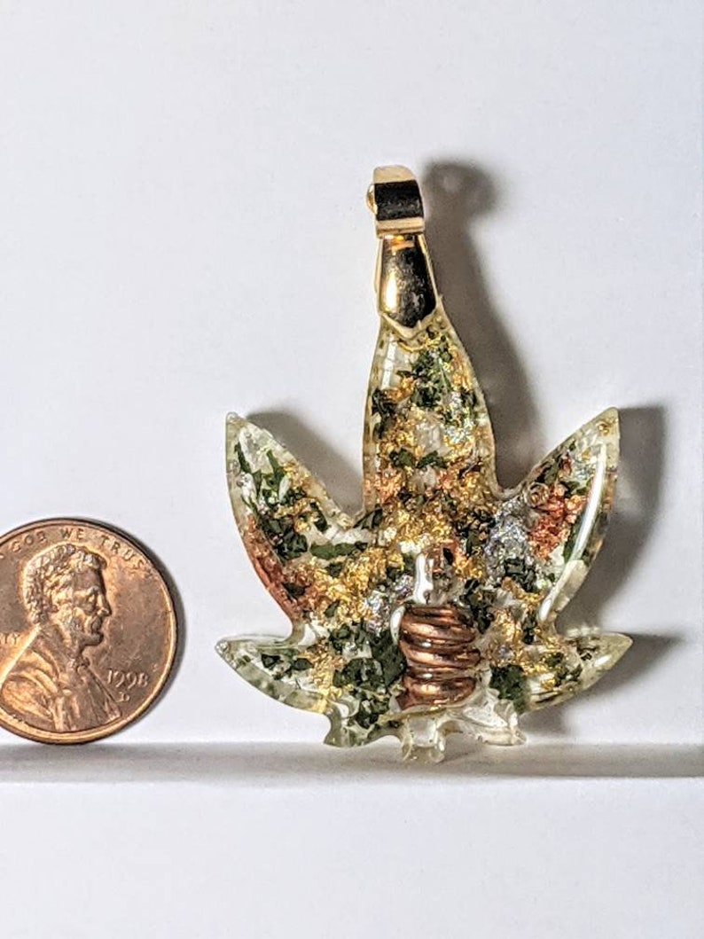 May include: Gold-tone pendant in the shape of a marijuana leaf with green, gold, and copper accents. The pendant is approximately the size of a US penny.