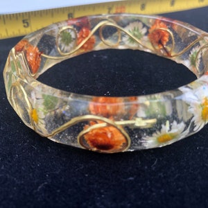 May include: A clear resin bangle bracelet with embedded dried flowers and gold wire. The flowers are white and orange.