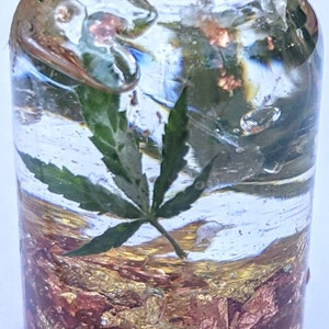 May include: A clear glass bottle filled with a golden liquid and gold flakes. A green marijuana leaf is suspended in the liquid.
